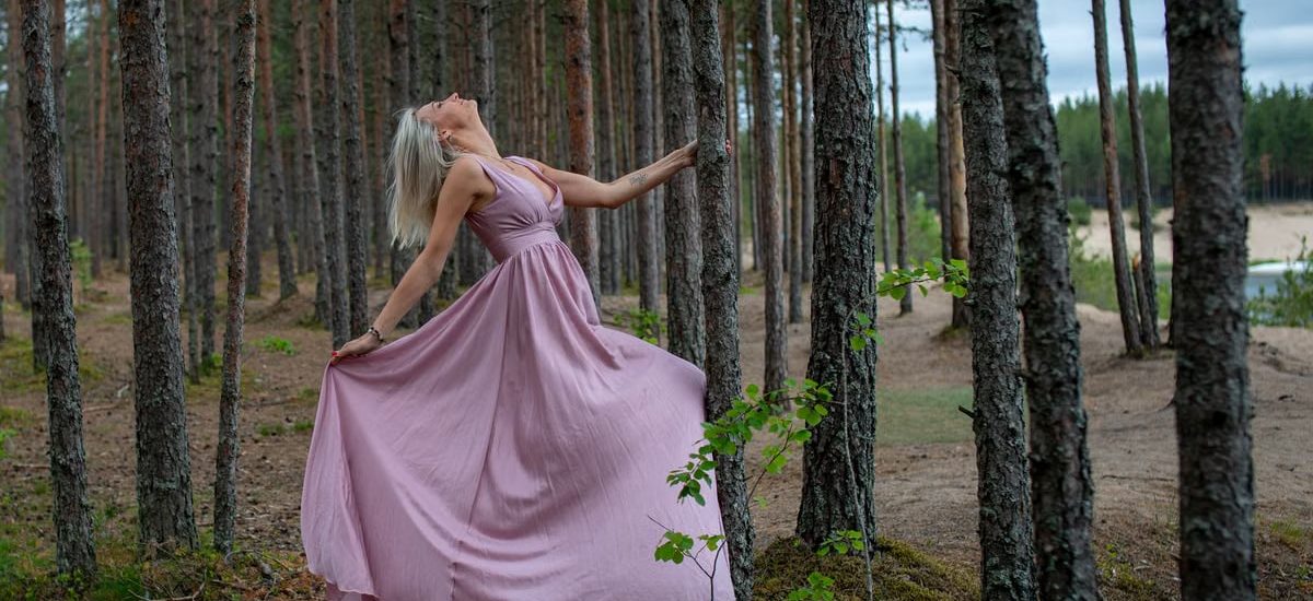 Blonde woman in flowing pink dress poses gracefully in Männiku forest, Estonia, during summer.
